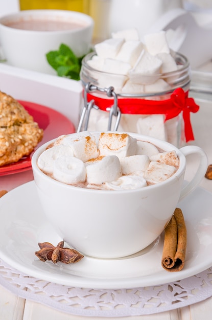 Taza de chocolate caliente caliente con malvaviscos caseros, canela y anís estrellado en una madera blanca.
