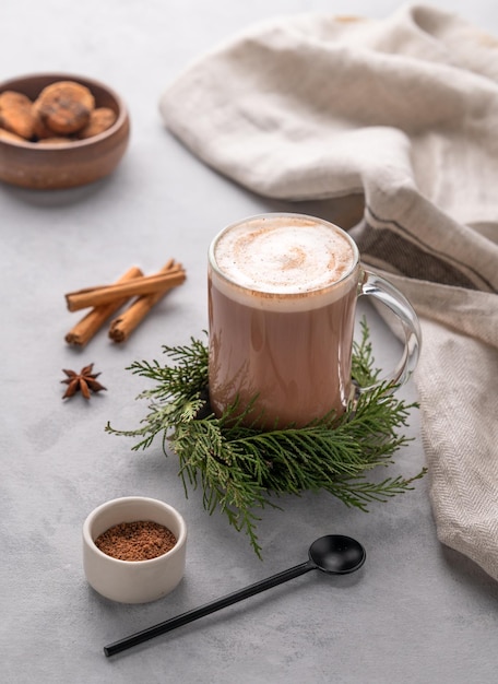 Taza de chocolate caliente con crema batida en un soporte con ramas de pino en un fondo claro con galletas de bastón de canela, cuchara de anís y polvo de cacao