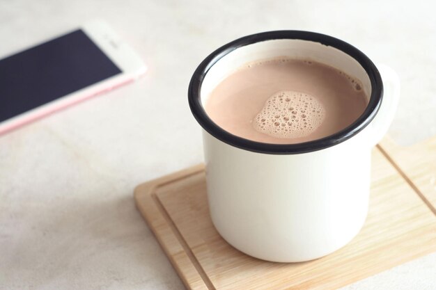 Una taza de chocolate caliente se encuentra sobre un soporte de madera sobre la mesa. Detrás hay un teléfono.