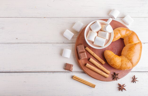 Una taza de chocolate caliente con malvavisco, croissant y especias sobre fondo blanco de madera.