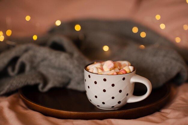 Taza de chocolate caliente con malvavisco con tejido de tela en bandeja de madera sobre luces de Navidad en la cama closeup Temporada de vacaciones de invierno