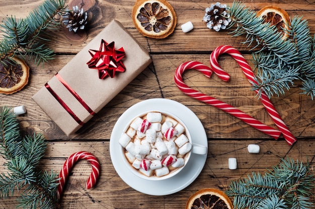 Taza de chocolate caliente con malvaviscos, caja de regalo, ramas de árboles de Navidad, bastones de caramelo y piñas