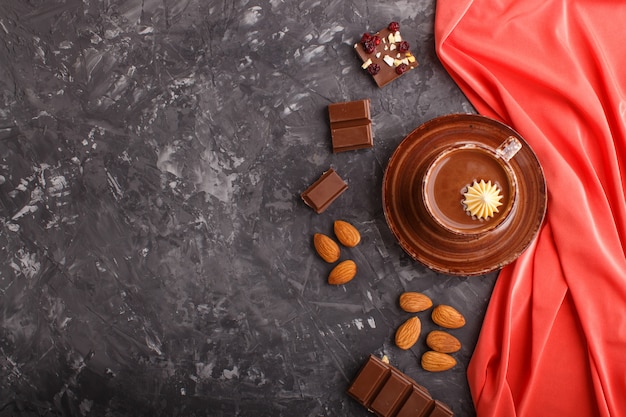 Taza de chocolate caliente y trozos de chocolate con leche con almendras sobre un fondo de hormigón negro con textil rojo.