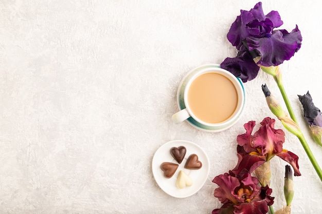 taza de cioffee con caramelos de chocolate y flores de iris morado y burdeos sobre fondo gris.