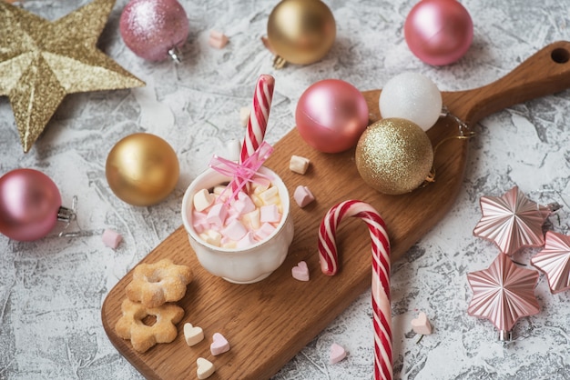 Taza de composición de Navidad o año nuevo con malvavisco en un soporte de madera con galletas y dulces entre juguetes brillantes de Año Nuevo sobre un fondo de textura brillante. Lay Flat, diseño