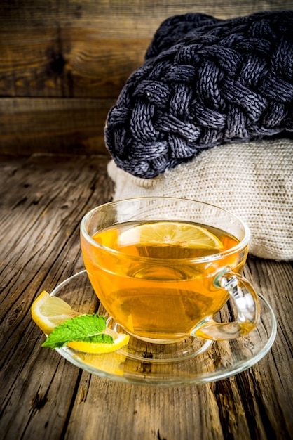 Taza de cristal de té con limón, menta y especias, en la vieja mesa de madera rústica con mantas calientes.