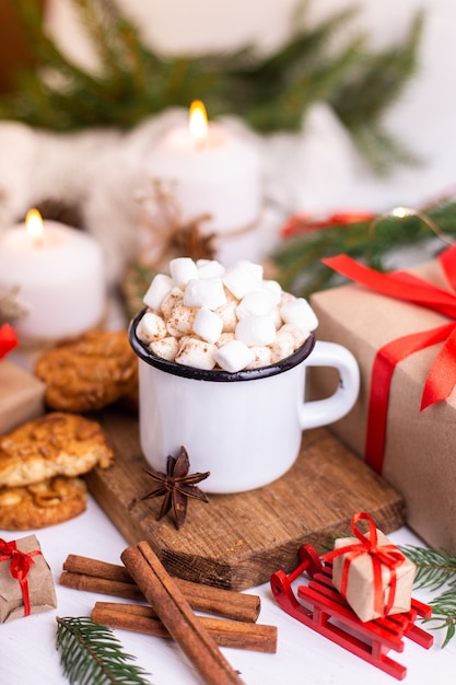 Taza esmaltada de chocolate caliente o café con malvaviscos y galletas. Alrededor de las ramas de los árboles, regalos y velas encendidas. Humor navideño. Postal o fondo de invierno.