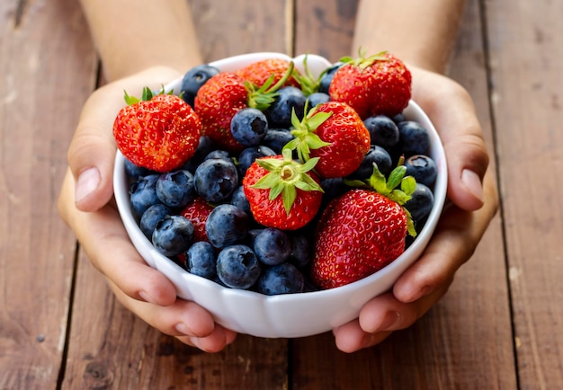 Una taza con fresas y arándanos maduros en manos de un niño al fondo de una mesa de madera