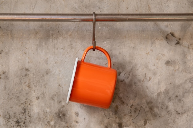 Taza de hojalata naranja colgada en riel inoxidable en pared de cemento