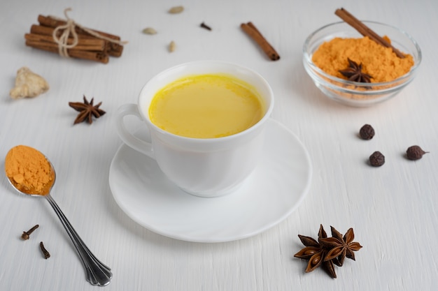 Taza de leche dorada servida en un plato con una cuchara de cúrcuma en polvo y canela en el cuadro blanco