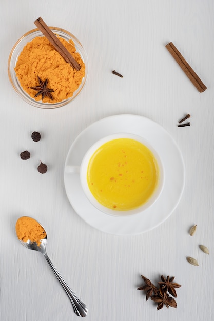 Taza de leche dorada servida en un plato con una cuchara de cúrcuma en polvo y canela en la mesa de madera blanca