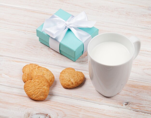 Taza de leche, galletas en forma de corazón y caja de regalo en la mesa de madera blanca