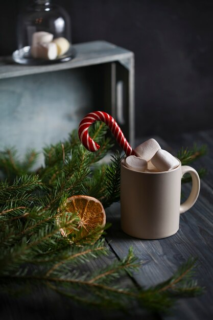 Taza ligera con cacao decorada con malvaviscos y bastones de caramelo, rodeada de ramas de abeto y una rodaja de naranja seca sobre un fondo de madera.