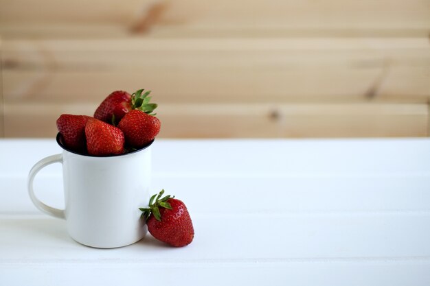 Una taza llena de fresas sobre un fondo de madera blanca