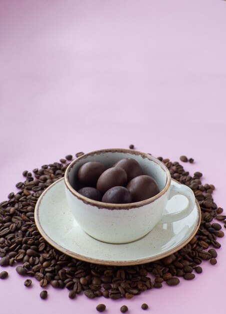 Taza llena de huevos de chocolate en un plato con granos de café sobre fondo rosa