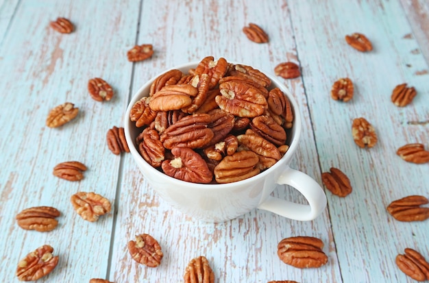 Taza llena de nueces pecanas con algunos granos esparcidos sobre una mesa de madera