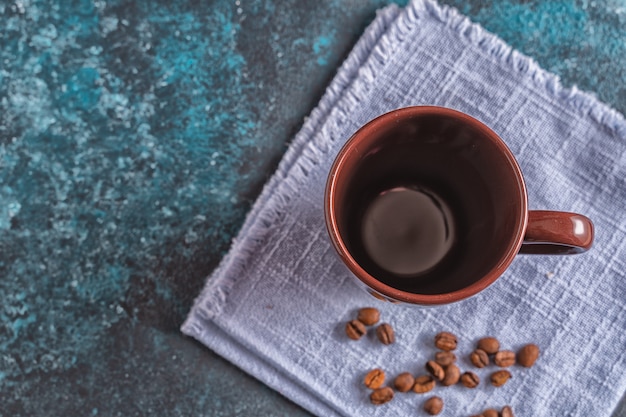 Taza marrón vacía para café y granos de café sobre fondo azul.