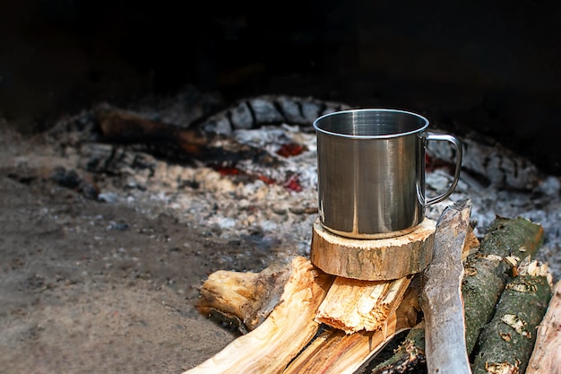 Una taza de metal para acampar se encuentra sobre leña junto al fuego Platos para acampar noche de fuego Copyspace