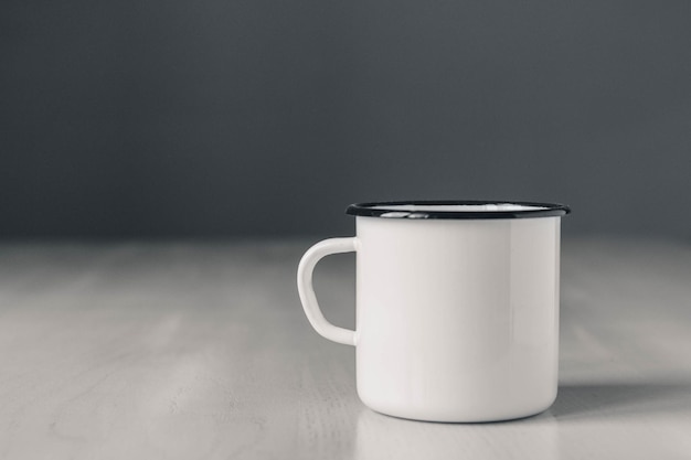 Taza de metal blanco esmaltado con línea negra en el borde de la mesa de madera blanca sobre fondo gris