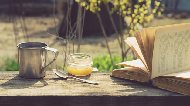 Taza de metal, un tarro de miel y un libro sobre una mesa de madera en el jardín