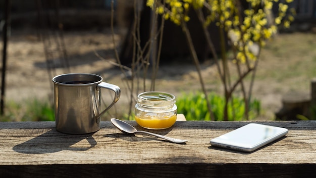 Taza de metal, un tarro de miel y un teléfono en una mesa de madera en el jardín