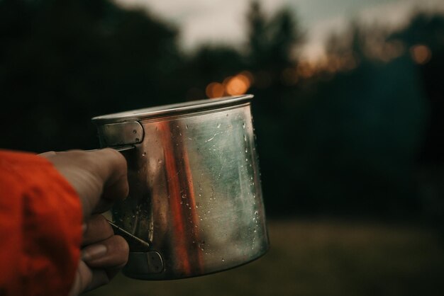 Taza de metal con té en la mano de un turista en el campamento de tiendas en las montañas bajo nubes blancas