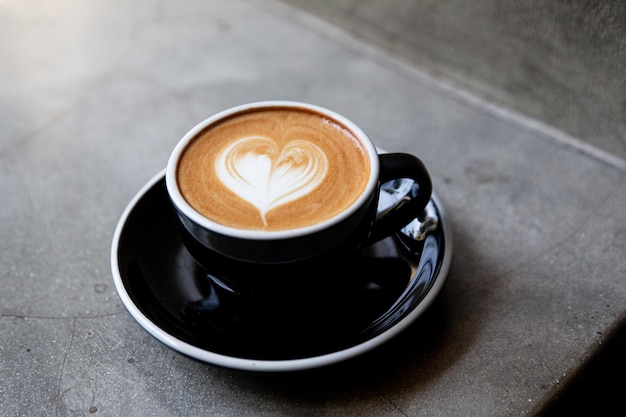 Taza negra de capuchino con arte latte de forma de corazón en platillo sobre fondo de hormigón