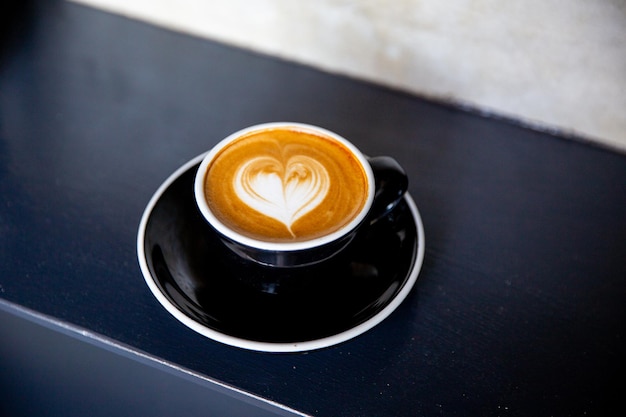 Taza negra de capuchino con arte latte de forma de corazón en platillo sobre fondo de hormigón