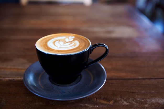 Taza negra de capuchino caliente en placa con textura azul sobre fondo de mesa de madera con arte latte.