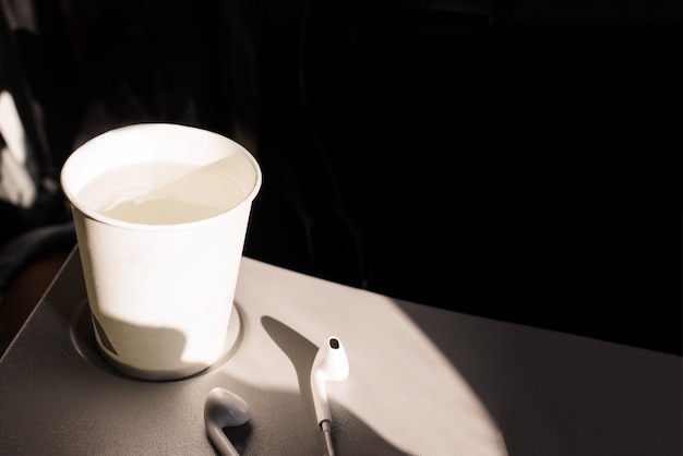 Taza de papel blanco de agua y auriculares en una mesa plegable en un avión. Bebidas en el avión. Deshidratación en vuelo.