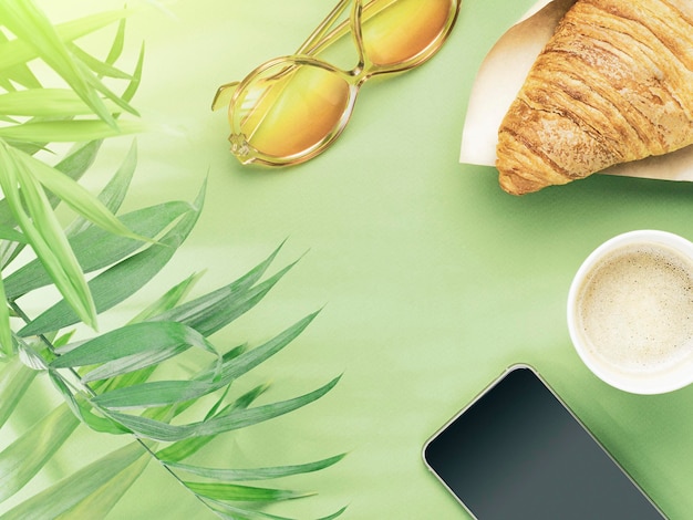 Taza de papel de café croissant teléfono gafas de sol y hojas de palma