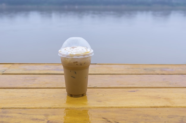 Taza plástica del café del capuchino de hielo en la tabla de madera al lado de la opinión del río