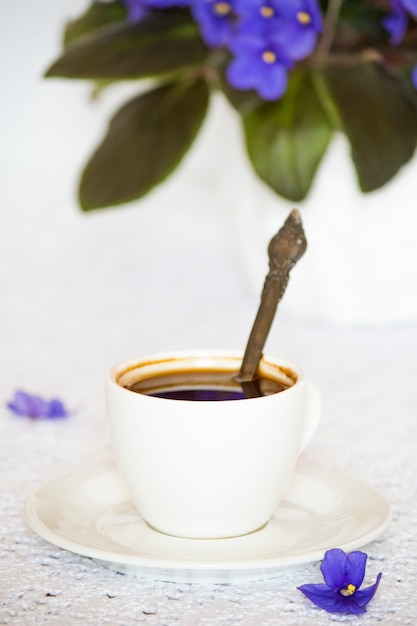 Taza y platillo de café con flores violetas africanas púrpuras sobre una superficie de mesa blanca.