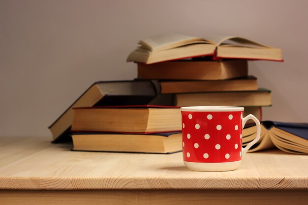 Taza de porcelana roja con lunares blancos y una pila de libros sobre una mesa de madera.