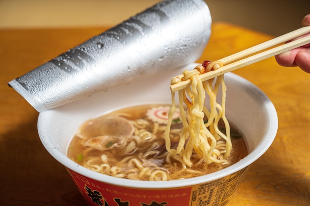 Una taza de ramen con palillos y una lata de agua.