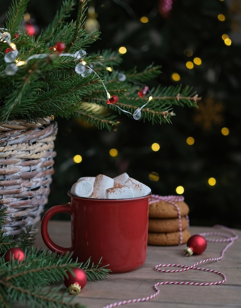 Taza roja de bebida caliente de invierno con malvaviscos y galletas en el fondo del árbol de Navidad