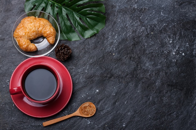 Foto taza roja de café y azúcar en una cuchara, croissant de pino seco sobre piedra negra