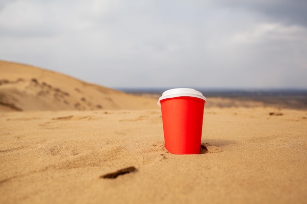 Una taza roja de café o una bebida caliente cubierta con una tapa en la arena del desierto Sandy seashore