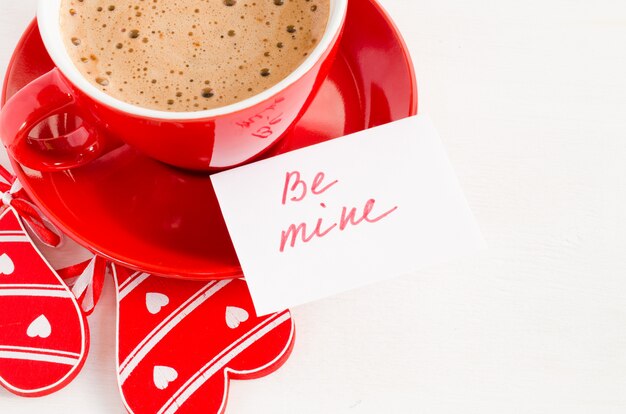 Una taza roja de capuchino con corazón de madera y notas Be Mine.