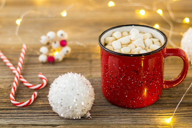 Taza roja de chocolate caliente con bolas blancas de malvaviscos y bastones de caramelo. fondo con hermosas luces de Navidad bokeh.
