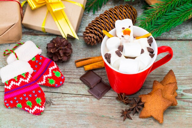 Taza roja con chocolate caliente con muñeco de nieve de malvavisco derretido sobre fondo de madera con regalos y adornos navideños.