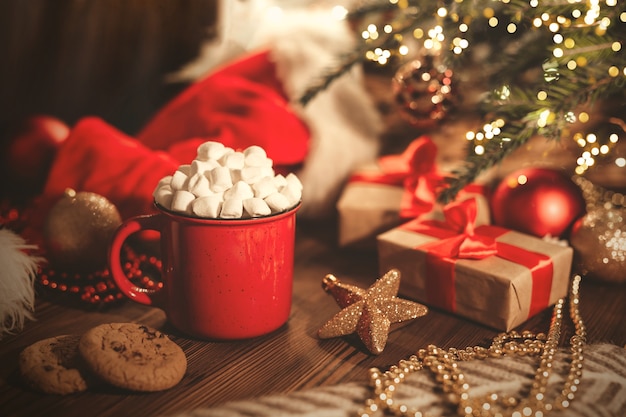Taza roja de Navidad con cacao y malvaviscos y galletas en una mesa de madera.