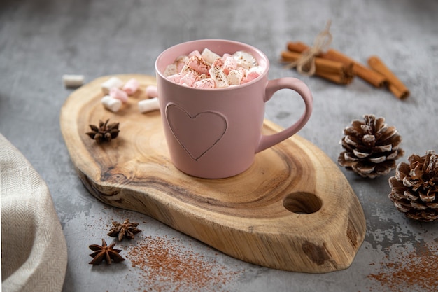 Taza rosa con cacao aromatizado y malvaviscos blanco-rosa, ramas de canela y anís estrellado sobre una tabla de madera
