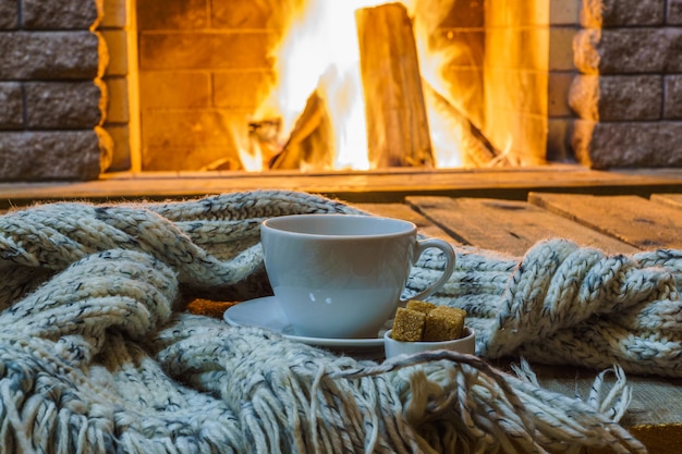 Taza de té y azúcar, cosas de lana cerca de una chimenea acogedora, en casa de campo, invierno
