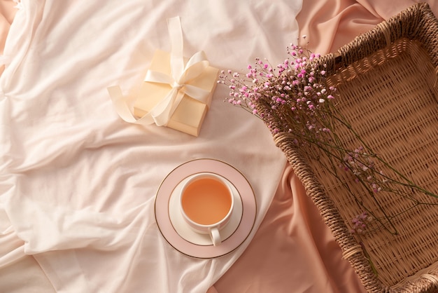 Taza de té, caja de regalo y flores en bandeja sobre fondo de tela ligera