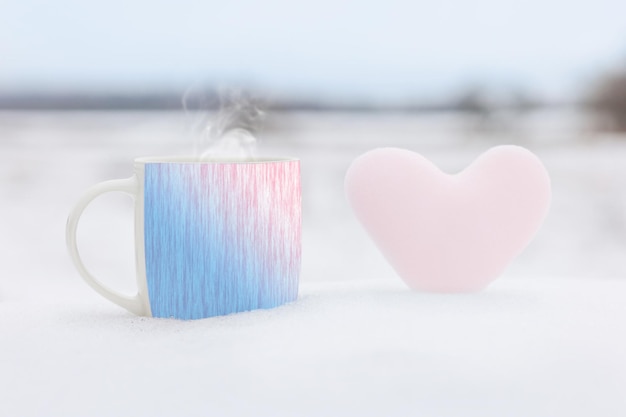 Una taza de té caliente con la imagen de liebres y un corazón rosa hecho de nieve en el fondo de un paisaje invernal Enfoque selectivo