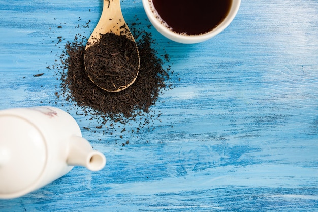 Taza con té caliente junto a una cuchara de madera con hojas de té sobre fondo azul vintage