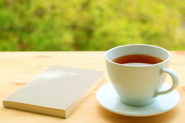 Taza de té caliente y un libro sobre una mesa al aire libre con follaje borroso en segundo plano.