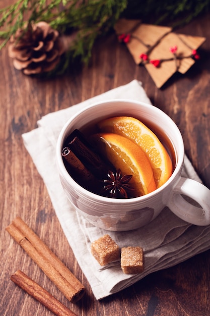 Taza de té caliente con naranja y especias sobre una rústica mesa marrón. De cerca