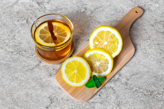 Una taza de té caliente con una rodaja de limón y menta para el tratamiento de la enfermedad en una tabla de cortar de madera sobre una mesa de piedra.